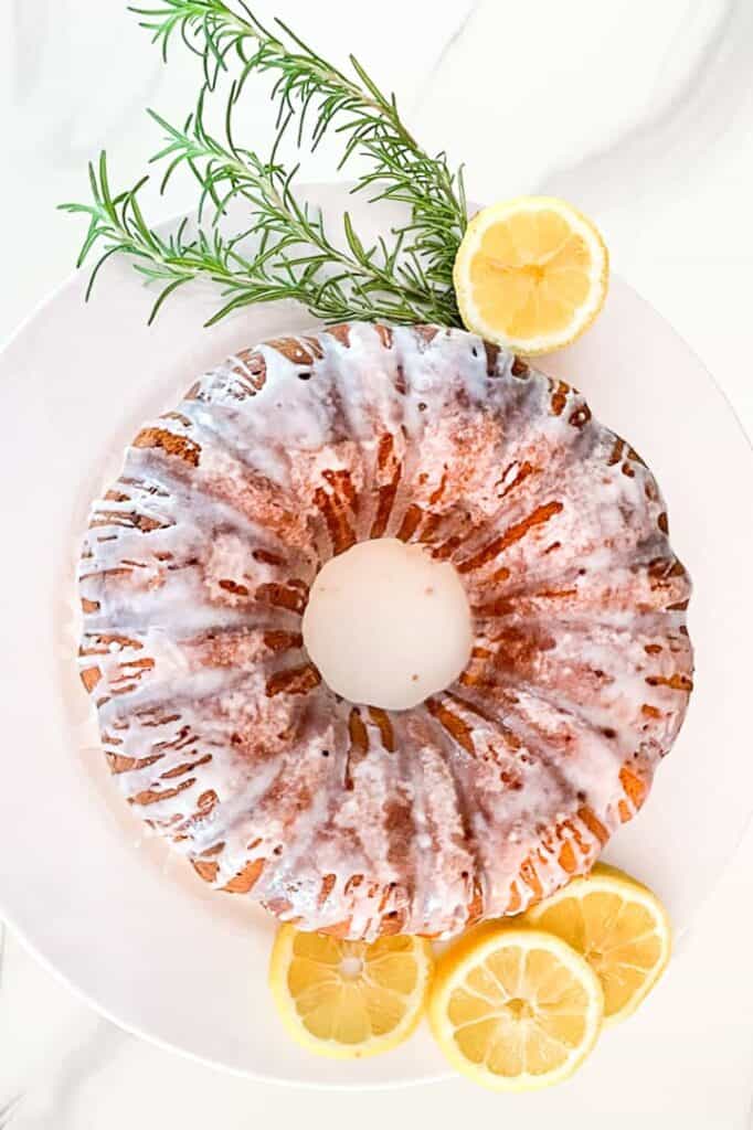 Lemon bundt cake sitting on a white plate with fresh lemon slices and rosemary. 