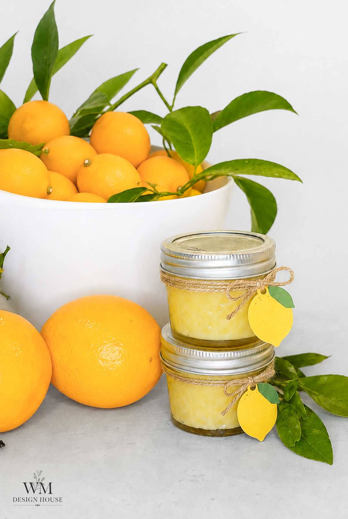 A white bowl filled with oranges and green leaves sits behind two small mason jars filled with a yellow spread, each tied with twine and a lemon-shaped tag—reminding you to always pick the good fruit, not bad lemons. Whole oranges and leafy branches rest nearby on a light surface.