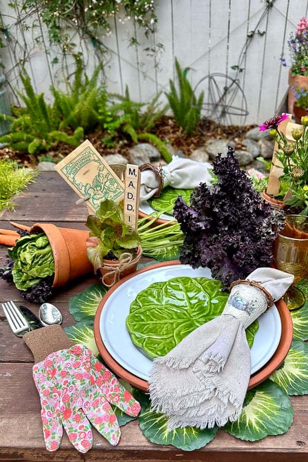A single place setting on the garden party table. The place setting starts with a cabbage placemat, a terra cotta charger, a white plate, and a small green cabbage plate. In addition, the table is decorated with terra cotta pots, lettuce, and a garden glove that houses the silverware. 