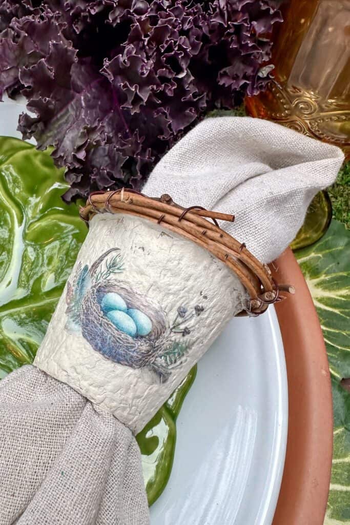 Peat pot napkin ring with the birds nest decal and a grapevine wreath around the edge that is holding a linen napkin. 