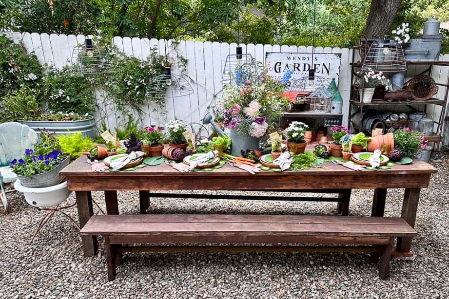 A large farm table is set with decorations for a garden party. The table is sitting on gravel in a garden yard with vintage decorations. Fresh flowers and vegetables adorn the table, which is surrounded by terra cotta pots and garden gloves. 