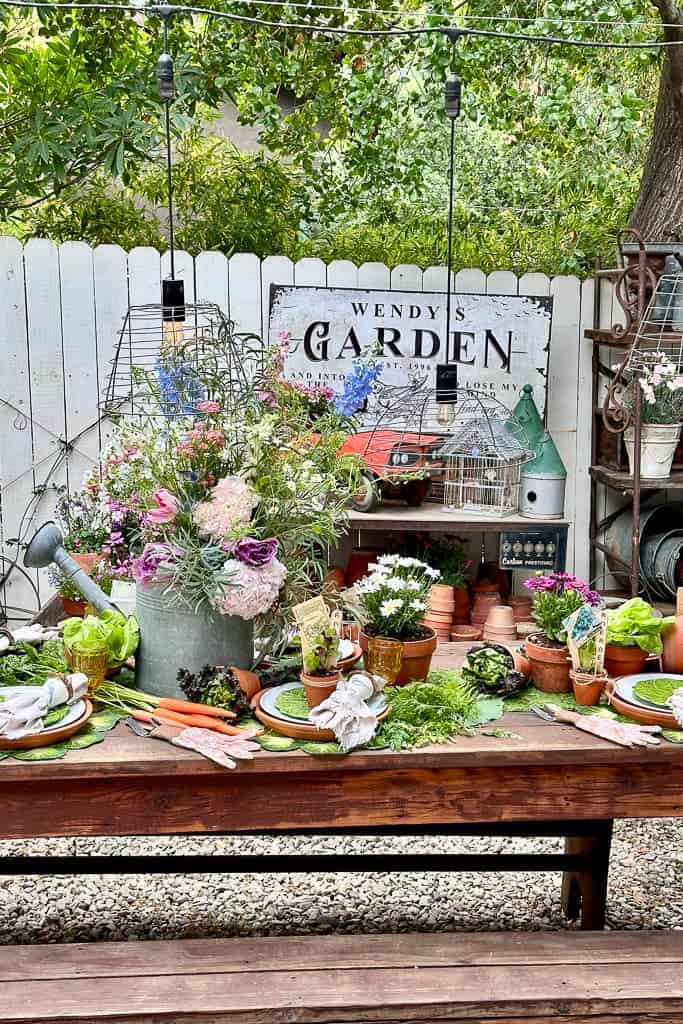 A centerpiece of fresh flowers housed in a galvanized watering can sits on a table set in the garden. 
