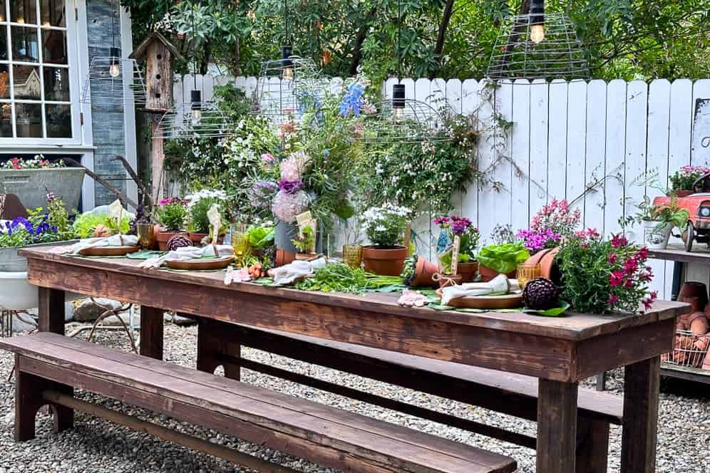 A large farm table is set with decorations for a garden party. Fresh flowers and vegetables adorn the table, which is surrounded by terra cotta pots and garden gloves. 