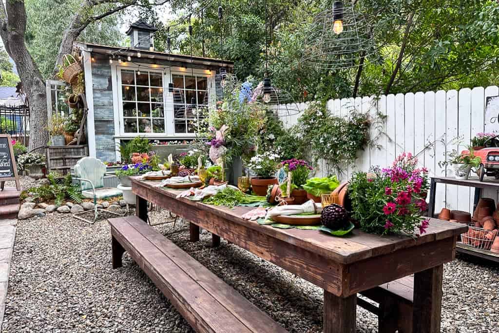 Garden Party Tablescape - A large farm table sits in a garden with a shed in the background. The table is set with garden decor of fresh flowers and vegetables.