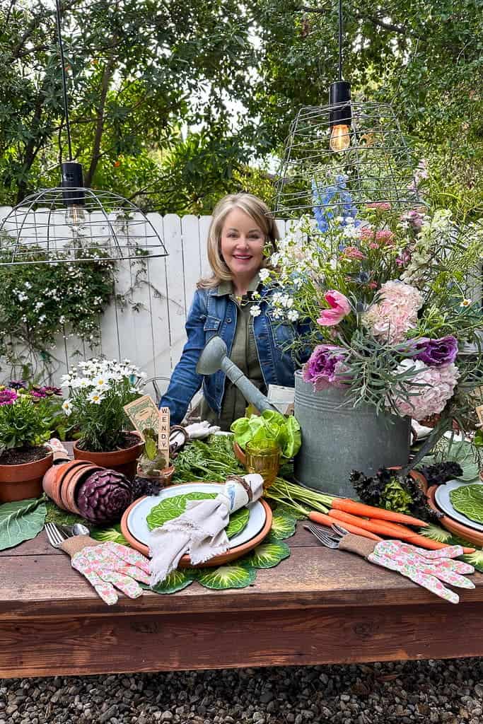 Garden Party Tablescape - A woman stands at a garden-themed table she designed for outdoor dining. A large watering can filled with spring flowers is the centerpiece, and the table is also decorated with loads of fresh vegetables. 