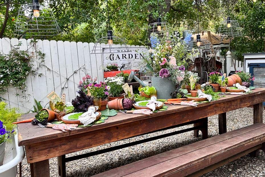 This is a garden party tablescape set in the garden. The table is decorated with clay pots, galvanized watering cans, fresh lettuce, carrots, artichokes, and fresh flowers. 