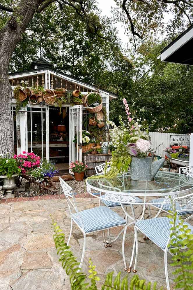 A shed decorated with baskets sits in a yard with a table, chairs, and beautiful flowers. 