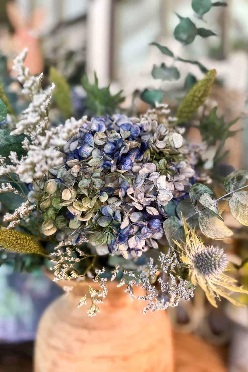 Close-up of a floral arrangement in a textured beige vase, featuring dried blue hydrangeas surrounded by various green leaves, white fillers, and a spiky silvery blue flower, set against a blurred natural background.
