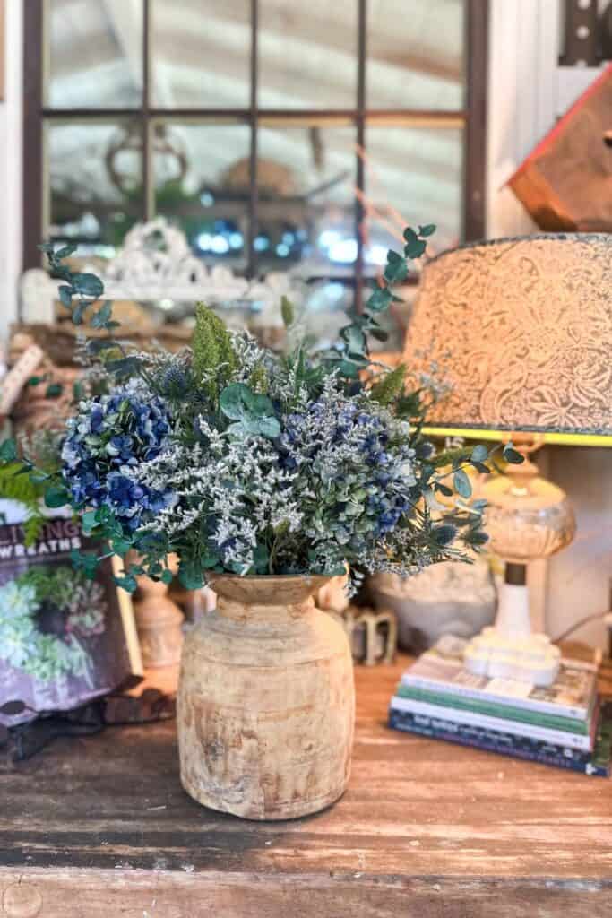 A rustic indoor setting featuring a large, earthy-toned vase filled with an arrangement of blue hydrangeas, white baby’s breath, and greenery. The vase sits on a wooden table next to a stack of books and a vintage-style lamp, with a window in the background.