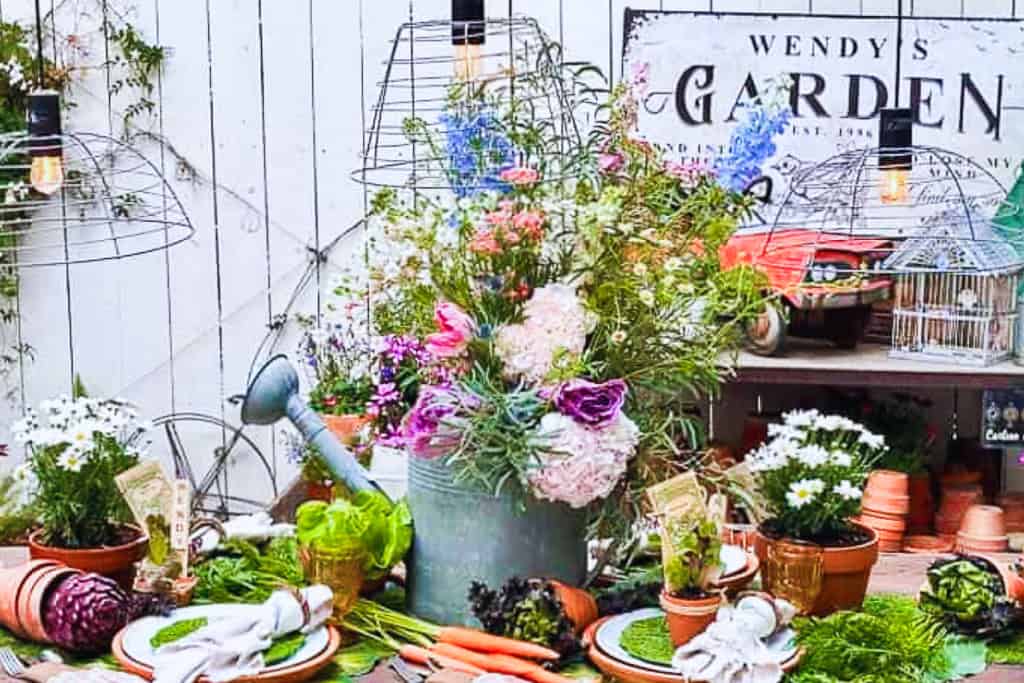 A rustic garden-themed table setting features a centerpiece with a watering can filled with vibrant flowers. The table is adorned with fresh vegetables, potted plants, garden tools, and charming birdcages. A sign reading "Wendy's Garden" hangs in the background.