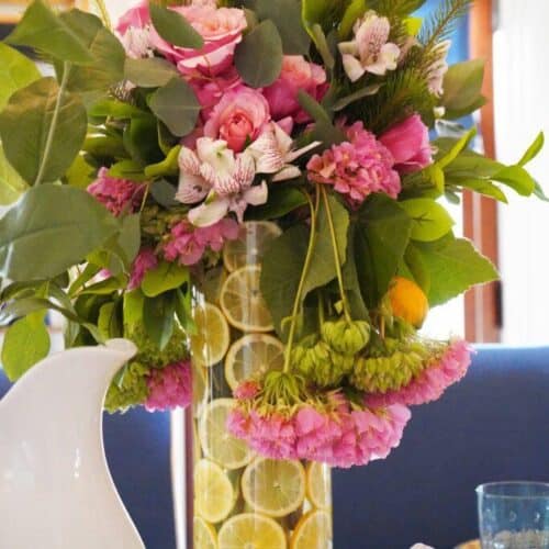 A floral arrangement featuring pink and white flowers with green foliage in a tall glass vase filled with lemon slices is a perfect spring decorating idea. The bouquet is placed on a white table next to a white pitcher and teapot, with a blurred blue and white background.