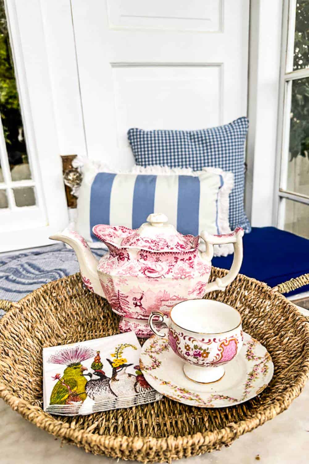 A wicker tray holds a pink and white porcelain teapot and a matching teacup with saucer. Next to the tea set is a decorative napkin featuring colorful birds. In the background, a window seat with blue and white cushions is visible against a white wall.