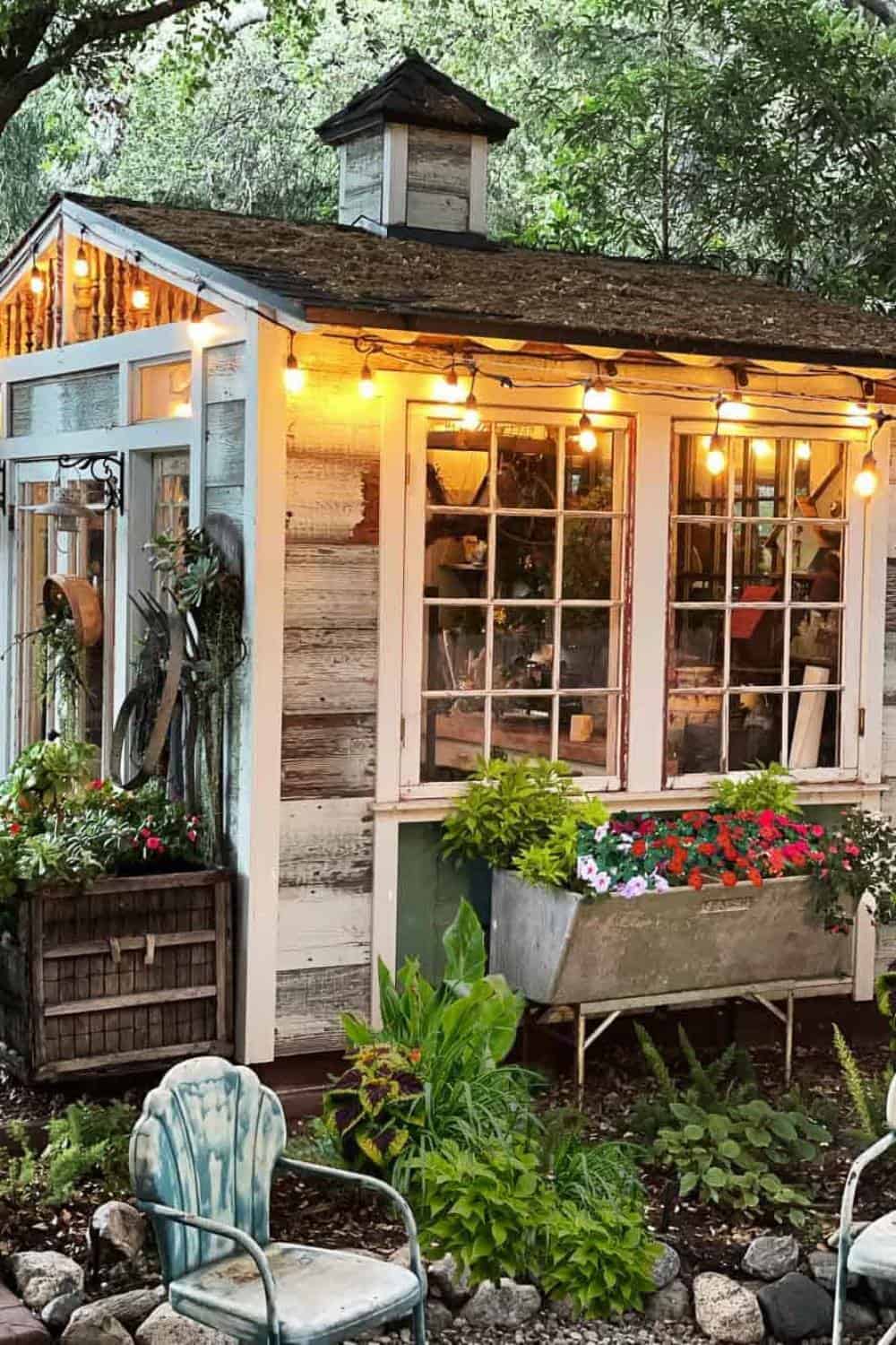 The She Shed lit up at night in the summer with string lights and flowers in planters around the outside