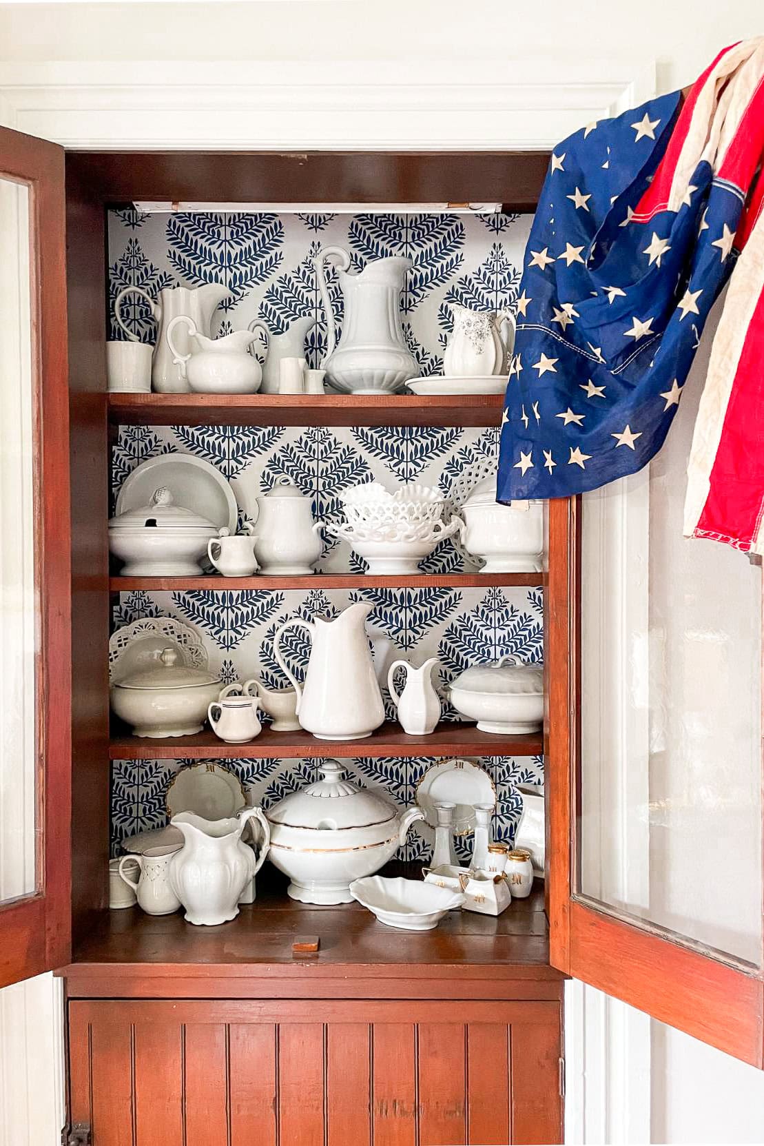 American  flag draped on the open door of a china hutch with a display of collected white vintage dishes
