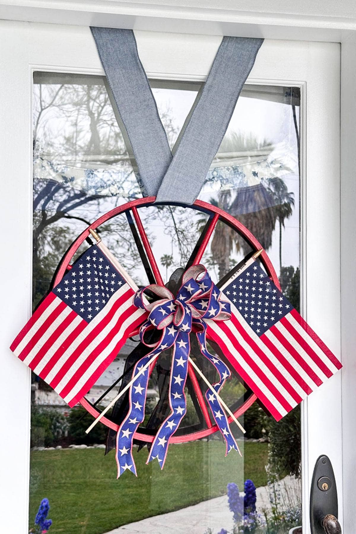 A circular door wreath adorned with two small American flags. The wreath is decorated with red, white, and blue ribbons featuring star patterns. It hangs using a wide denim-like fabric and is placed against a glass door with an outdoor scene visible through it.