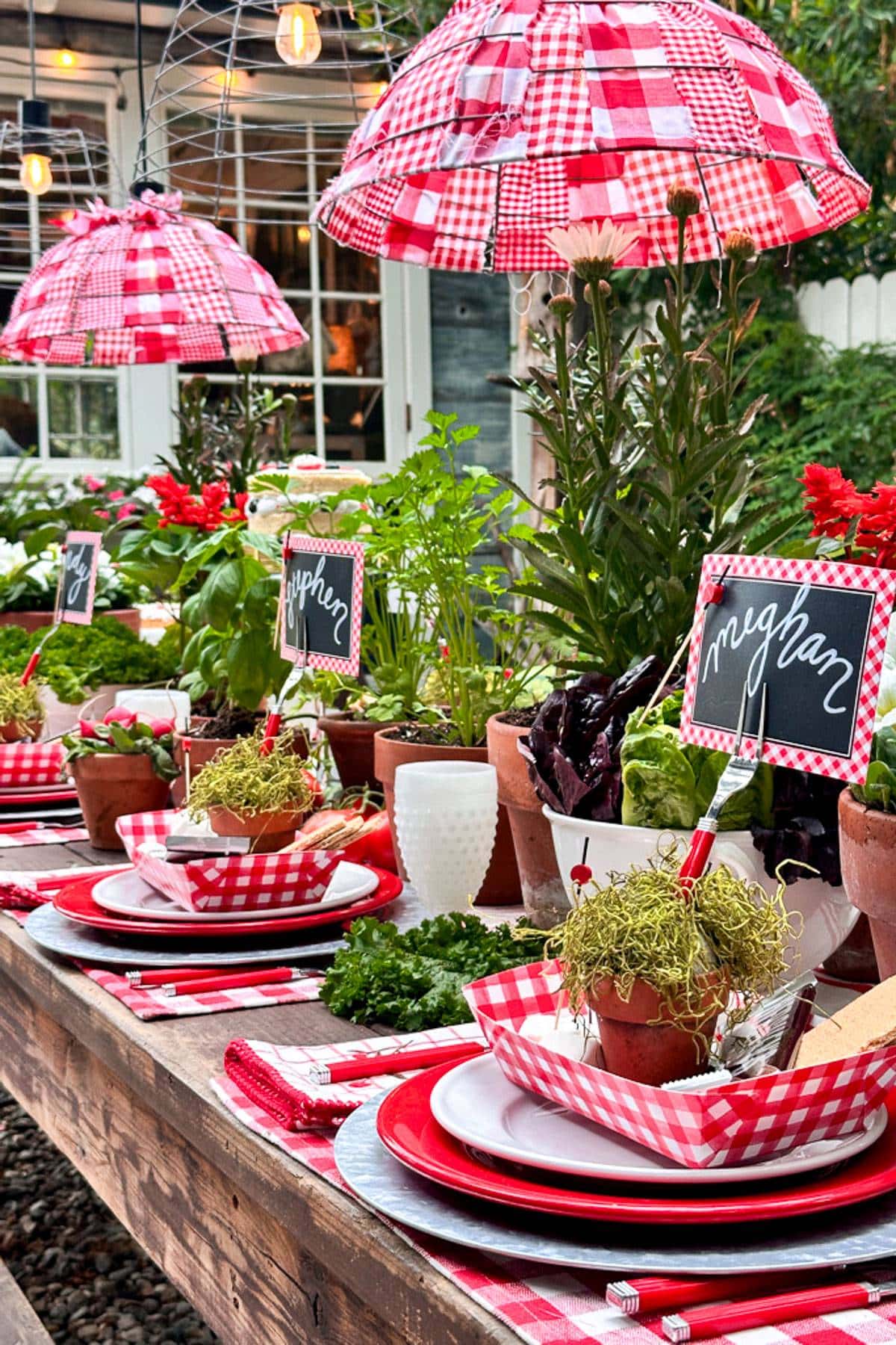 A farm table set for a smokin' hot summer tablescape. Red and white check light fixtures hang over a table of herbs and plants in terra cotta pots. Red and white check placemats, red and white dishes, and fun place cards sit in forks. 