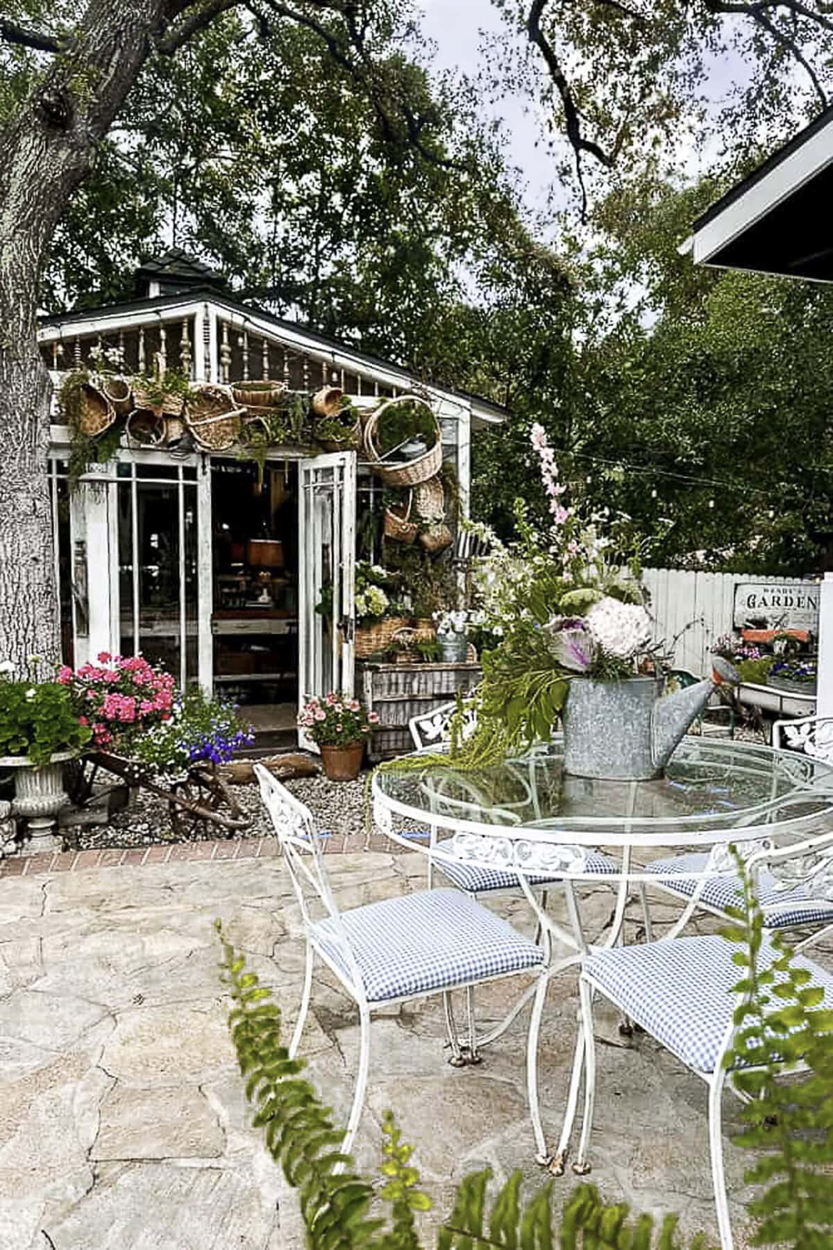 
A she shed decked out for the summer, featuring an arch of baskets, sits next to a patio with a vintage table and chairs, perfectly set for a summer luncheon.