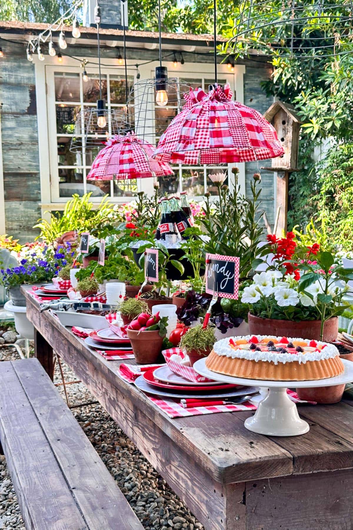 Summer tablescape in red and white colors. Two lampshades adorn the table with red and white check, and fruits and vegetables with fresh herb plants decorate the table. 