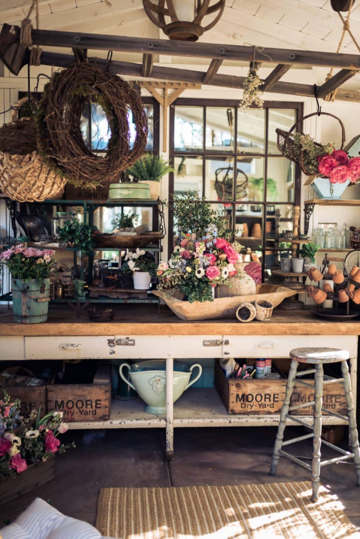 A cozy potting shed features a wooden workbench adorned with various flowers and gardening tools. Hanging baskets, potted plants, and a rustic stool add charm. Large mirrors reflect the bright, inviting space filled with boxes labeled "MOORE's Dry Yard.