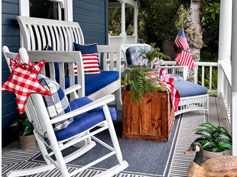 Front Porch decorated for the fourth of July with a rocking chair, a bench and a vintage box for a coffee table. a blue rug and blue and white cushions.
