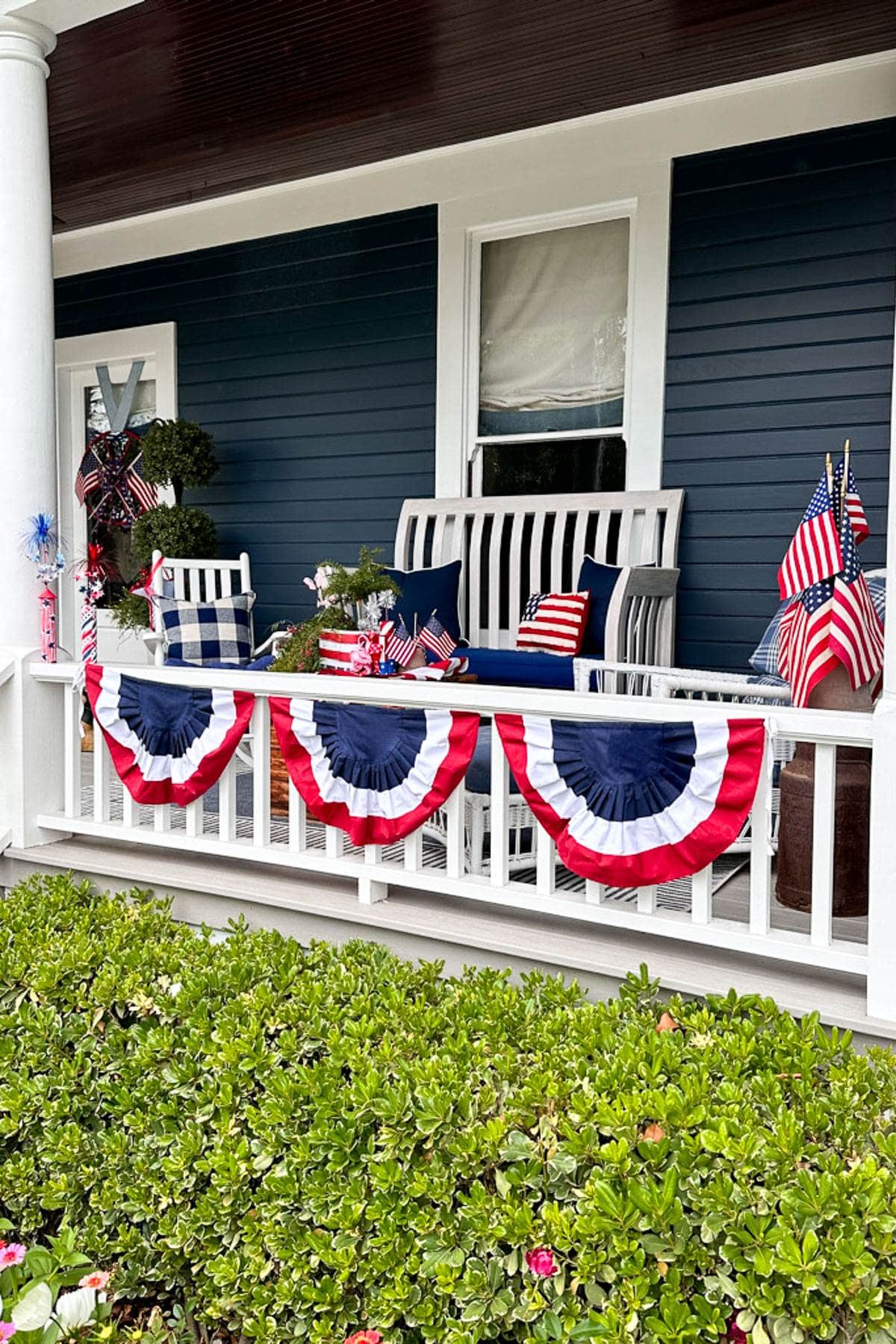 A porch decorated for a patriotic celebration: red, white, and blue bunting hangs from the railing, a cluster of American flags is displayed, and two chairs with cushions sit near a small table adorned with festive decorations, including more American flags. Green shrubs line the foreground.