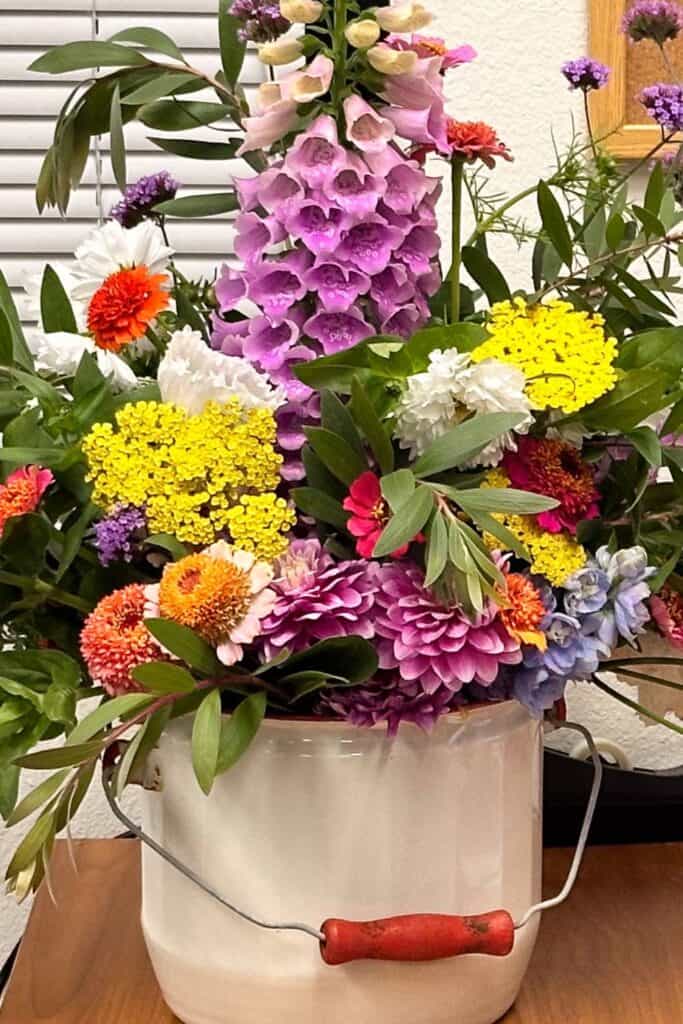 A colorful bouquet of various flowers, including pink, yellow, and orange blossoms, arranged in a white metal bucket with a red handle. The bucket sits on a wooden surface, with greenery and a white wall with blinds in the background.
