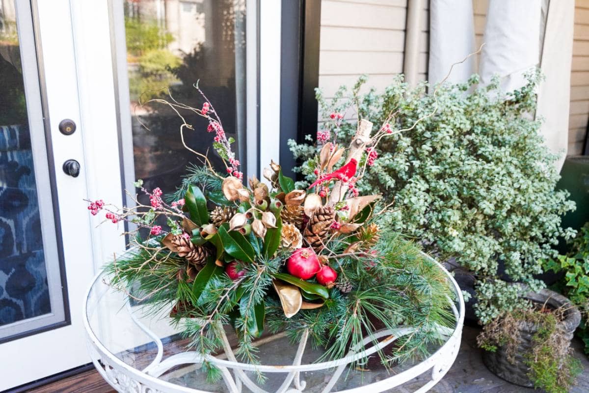 A festive outdoor arrangement on a round white table features pine branches, pine cones, red berries, and glossy dark green leaves. Decorative golden leaves and red apple ornaments add a touch of holiday glamour. A green leafy plant can be seen in the background.