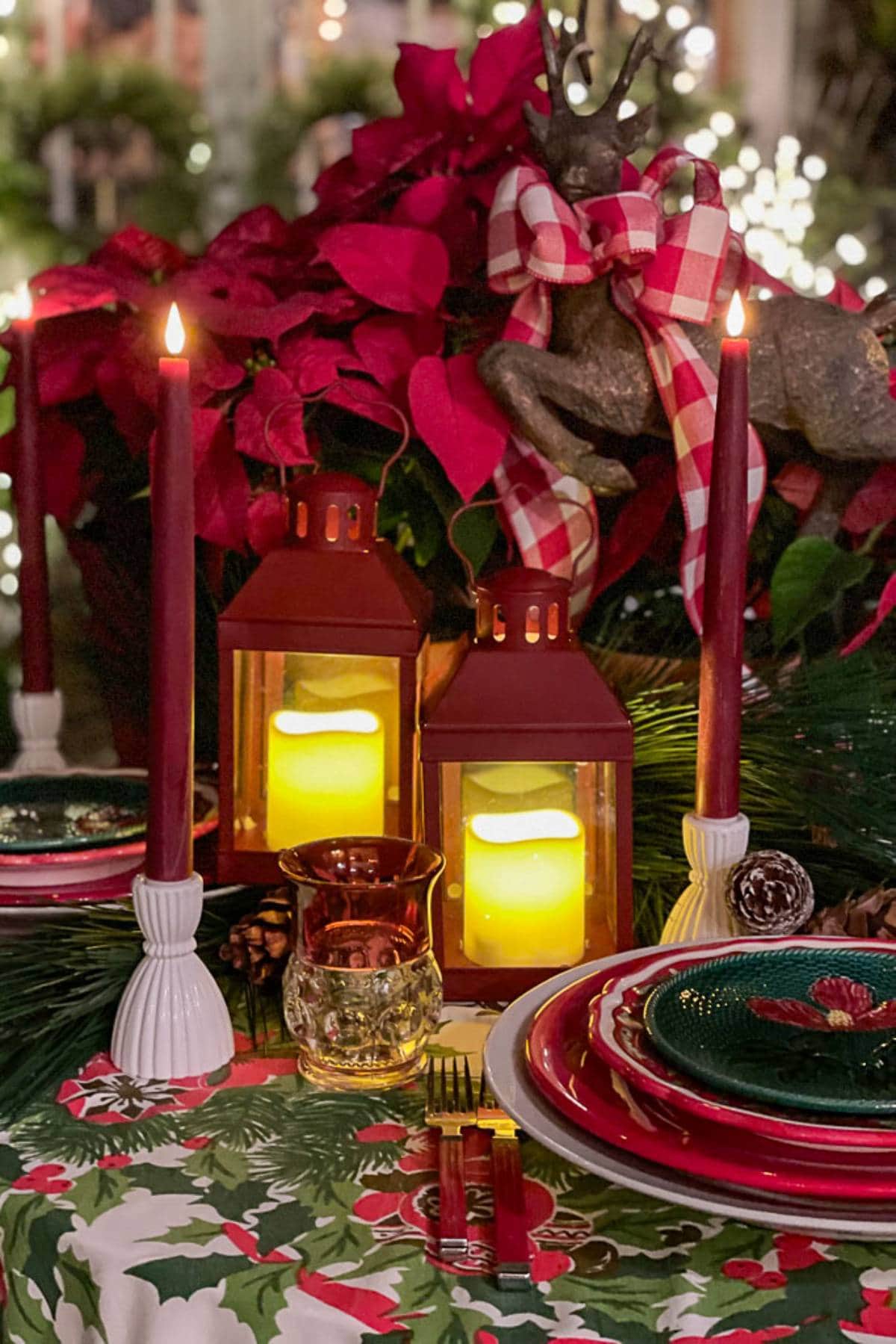 A festive Christmas table setup with red and green decor. Two lit red candles flank two glowing lanterns in the center. The table is adorned with poinsettias, pine leaves, and a reindeer figure draped in a checkered ribbon. Plates are layered with holiday designs.