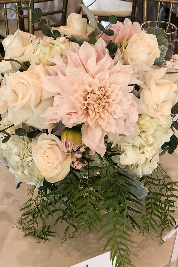 A floral arrangement featuring a large pale pink dahlia at the center, surrounded by soft white roses, cream-colored hydrangeas, and delicate greenery, displayed in a translucent vase on a light-colored tablecloth—an ideal option for outdoor centerpiece ideas.