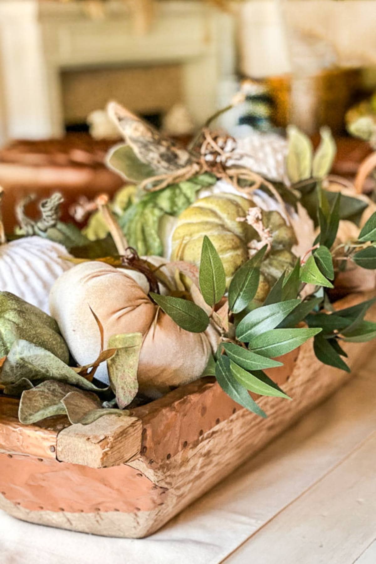 A wooden rectangular container is filled with various decorative pumpkins and green foliage, creating an autumn-themed centerpiece on a light-colored tablecloth. The background shows a slightly blurred fireplace.