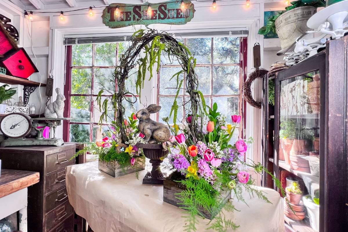 A room adorned with vibrant spring flowers, including tulips and daisies. A decorative rabbit figurine sits in the center of a table under a rustic arch made of branches and greenery. Various gardening items and potted plants surround the area near a large window.