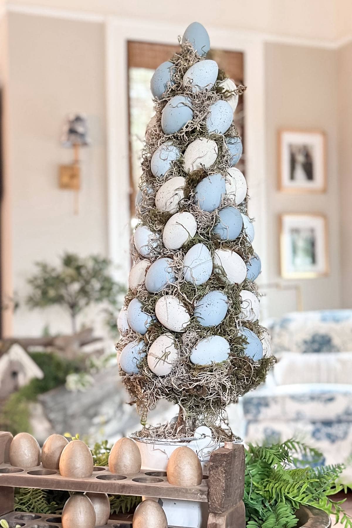 A decorative tree adorned with blue and white speckled eggs stands on a table in a room. Moss weaves through the eggs, creating a natural look. Nearby is a tray with smaller golden eggs. The scene is set indoors with plants and framed pictures in the background.