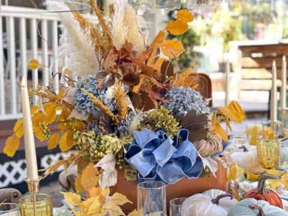 A festive autumn centerpiece featuring a mix of dried leaves, pampas grass, and blue hydrangeas arranged in a vase. The table is set with glassware, candles, and decorative pumpkins. One of the best outdoor centerpiece ideas, it enhances a porch adorned with white railings and outdoor furniture.