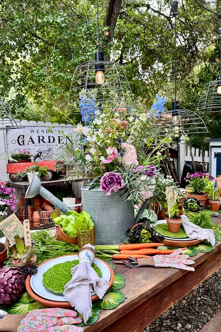 A beautifully set outdoor garden dining table with floral arrangements in a watering can, potted plants, gardening tools, and vibrant place settings. Overhead pendant lights hang from above a sign reading "Garden," offering perfect outdoor table centerpiece ideas.