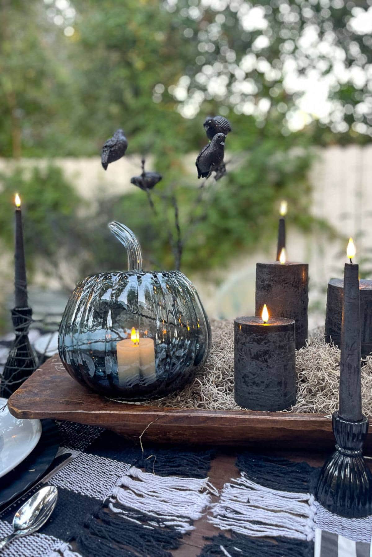 An outdoor Halloween-themed table setting features dark candles, a blue glass pumpkin with a lit candle inside, and black crow decorations. The table is adorned with moss and black and white placemats, with a natural, blurred background of greenery.