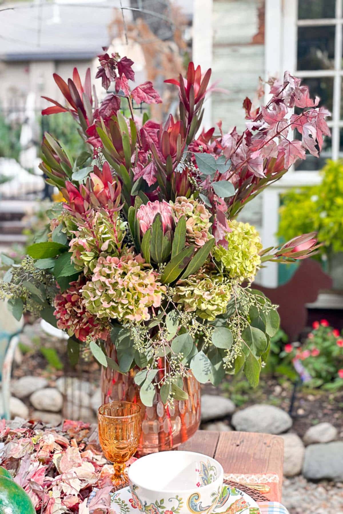A vibrant floral arrangement sits in a copper vase on an outdoor table. The bouquet includes green and dusty pink hydrangeas, leafy branches, and dark red foliage. Nearby are an orange glass, a white bowl with a colorful pattern, and fallen autumn leaves.