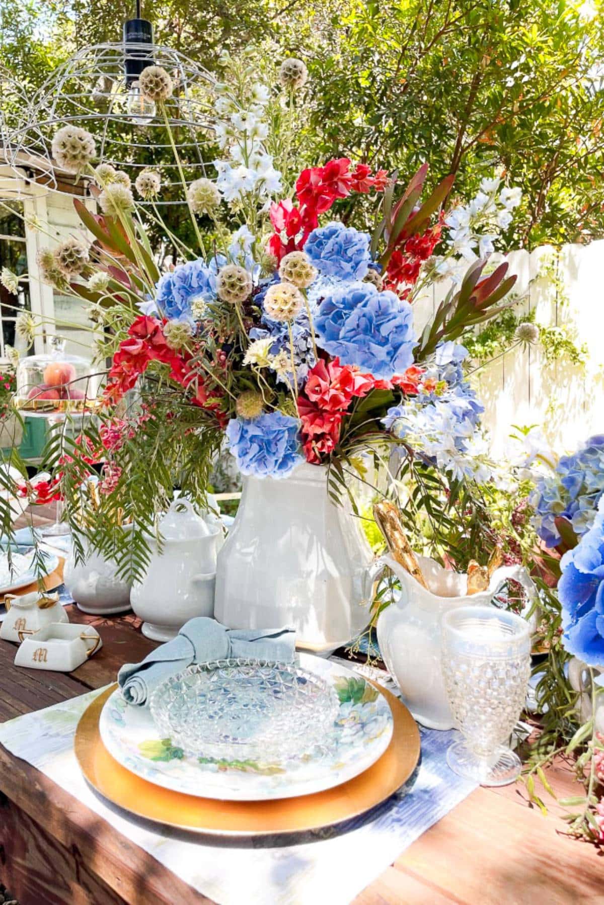 A vibrant outdoor table setting featuring a large white pitcher filled with blue, red, and white flowers as the centerpiece. Surrounding the pitcher are white teapots, a decorative drink dispenser, clear glassware, and a plate on a gold charger. Greenery in the background.