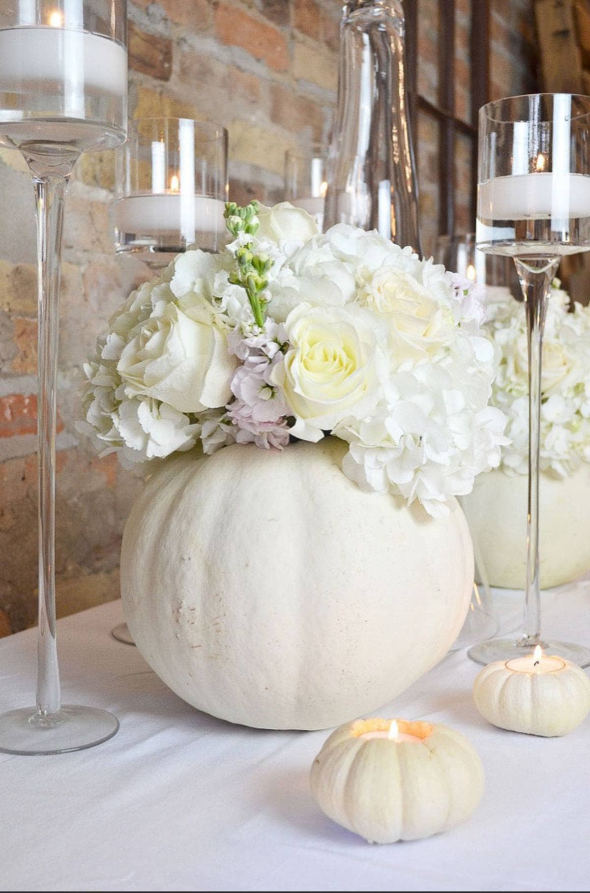 A decorative fall centerpiece features a white pumpkin filled with a bouquet of white flowers, including roses and hydrangeas, placed on a table. Tall glass candle holders with lit candles and small white pumpkins with candles are arranged around it.