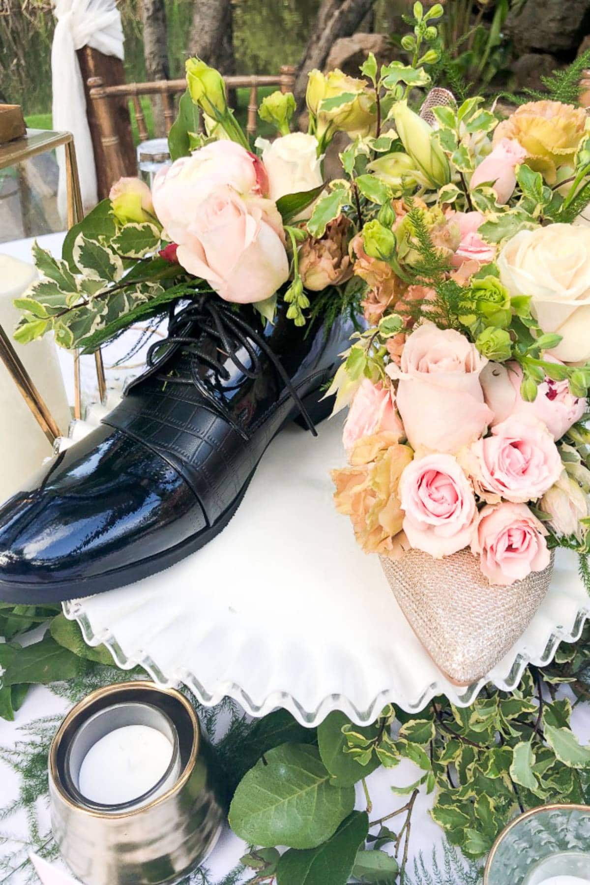 A polished black dress shoe and a glittery high heel filled with flowers are displayed on a white ruffled plate, surrounded by pink and creamy white roses. A candle in a metallic holder is placed nearby, along with various green foliage and decorative elements.