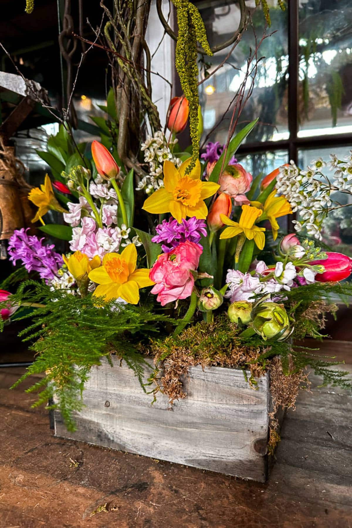 A wooden box filled with a vibrant arrangement of various spring flowers, including yellow daffodils, pink tulips, purple hyacinths, and greenery, sits on a wooden surface near a window. The lush bouquet exudes freshness and color, creating a cheerful display.