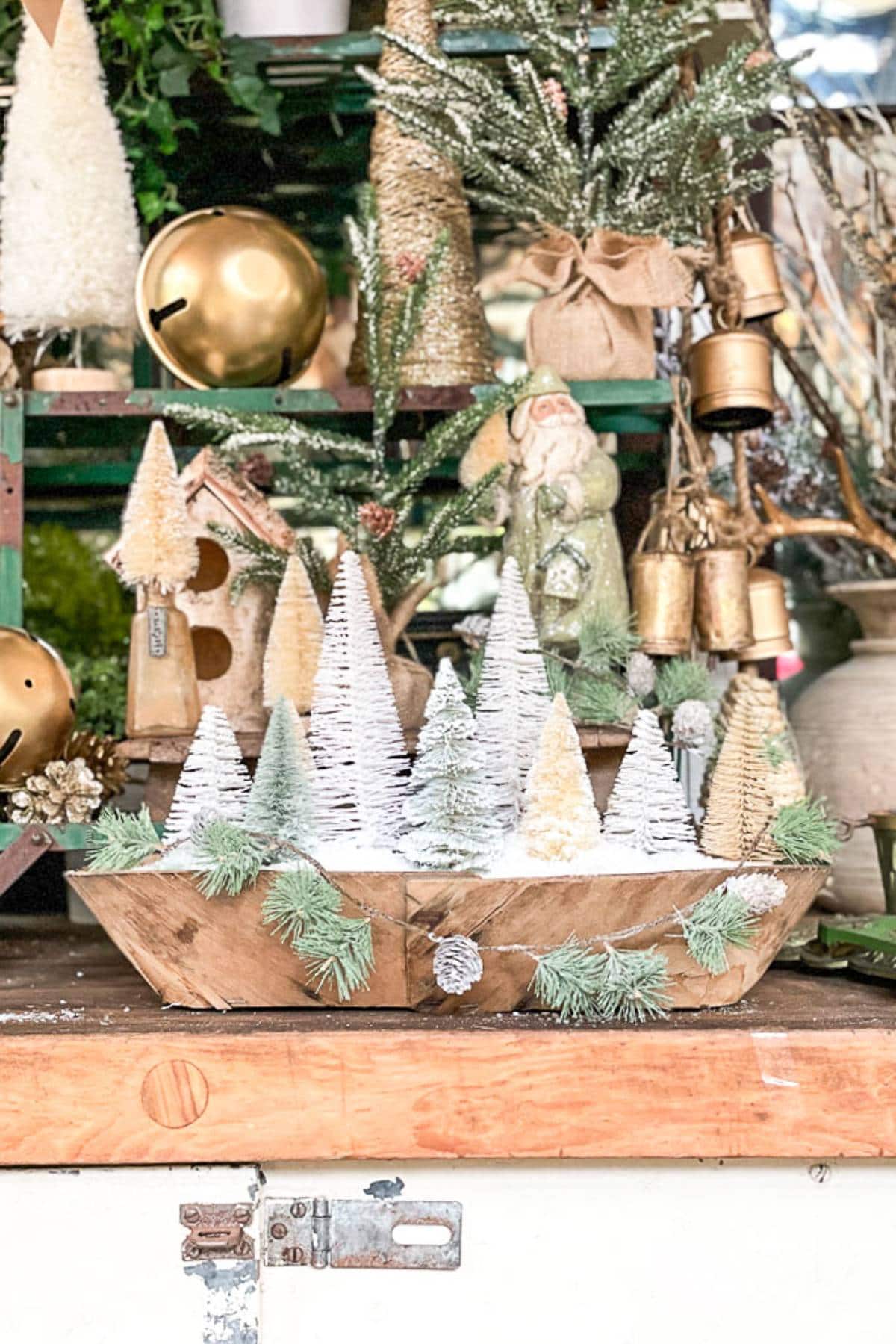 A festive holiday display featuring a wooden bowl filled with small, snow-covered, artificial Christmas trees. Surrounding the bowl are other holiday decorations such as bells, a birdhouse, and a figurine of Santa Claus, all accented with greenery and ornaments.