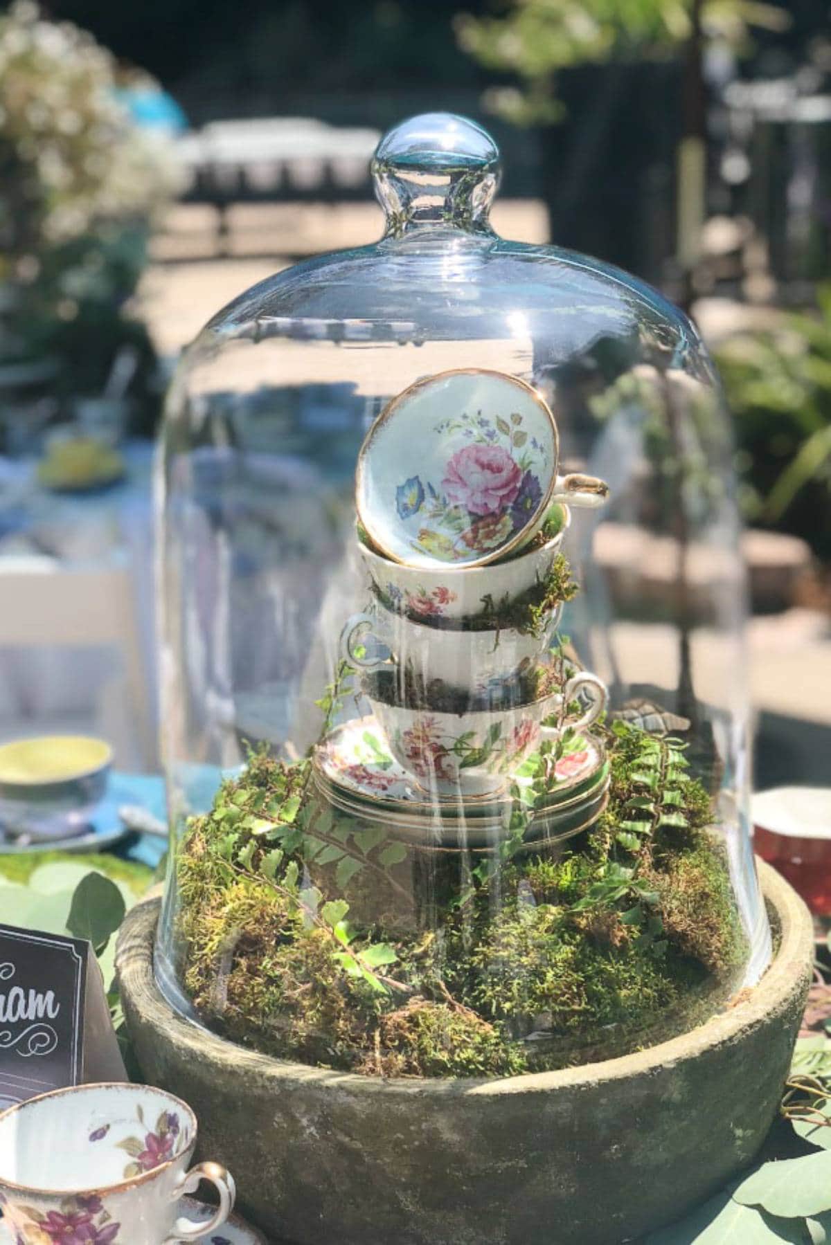 A stack of floral-patterned teacups and saucers is displayed under a glass cloche on a moss-covered base. The setup is part of an outdoor tea party with various other teacups and greenery visible in the background.