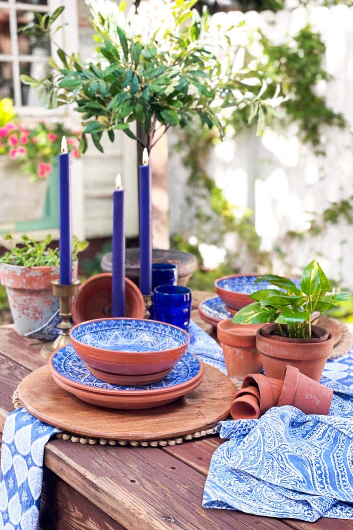 A rustic outdoor table is set with blue-patterned bowls, terra cotta plates, and small potted plants. Blue candles in wooden holders are lit, and the table is adorned with a blue patterned cloth. Greenery and a window with hanging flowers are in the background.