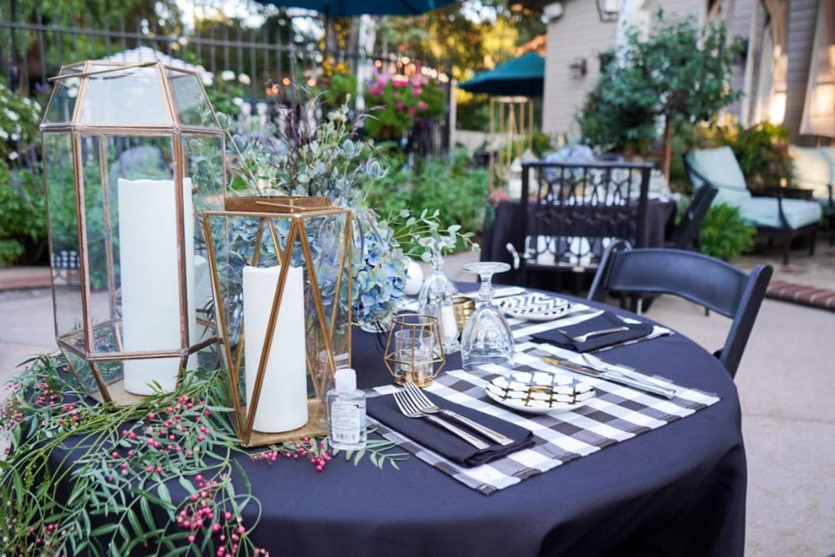 A round table set for an outdoor event with black tablecloth, black and white checkered napkins, and gold-accented plates and glasses. Candle holders, greenery, and flowers decorate the table. Surrounding area includes a garden, a fence, and patio furniture.