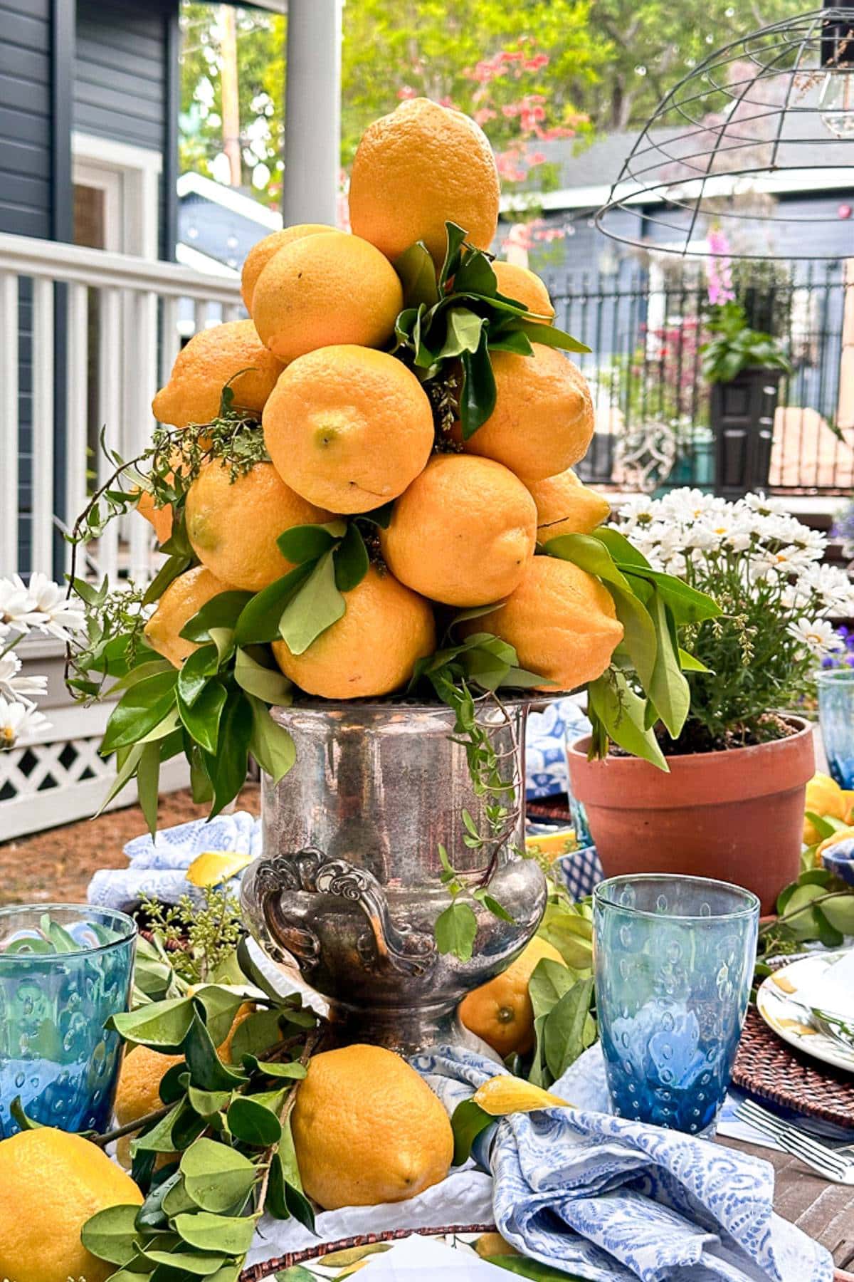 A decorative arrangement of fresh lemons and green leaves piled high in a silver urn centerpiece, surrounded by blue-tinted glasses, white flowers in a terracotta pot, and folded napkins with blue patterns on an outdoor table.