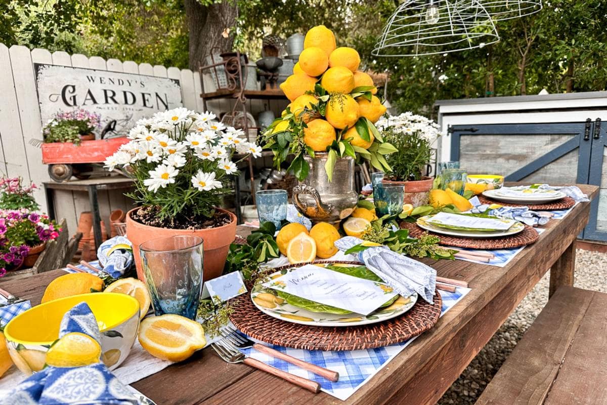 A rustic outdoor table setting adorned with lemons and white flowers. Round wicker placemats hold white plates with lemons and printed menus. A silver pitcher filled with lemons is a centerpiece. "Wendy's Garden" sign and greenery in the background complete the garden-themed ambiance.