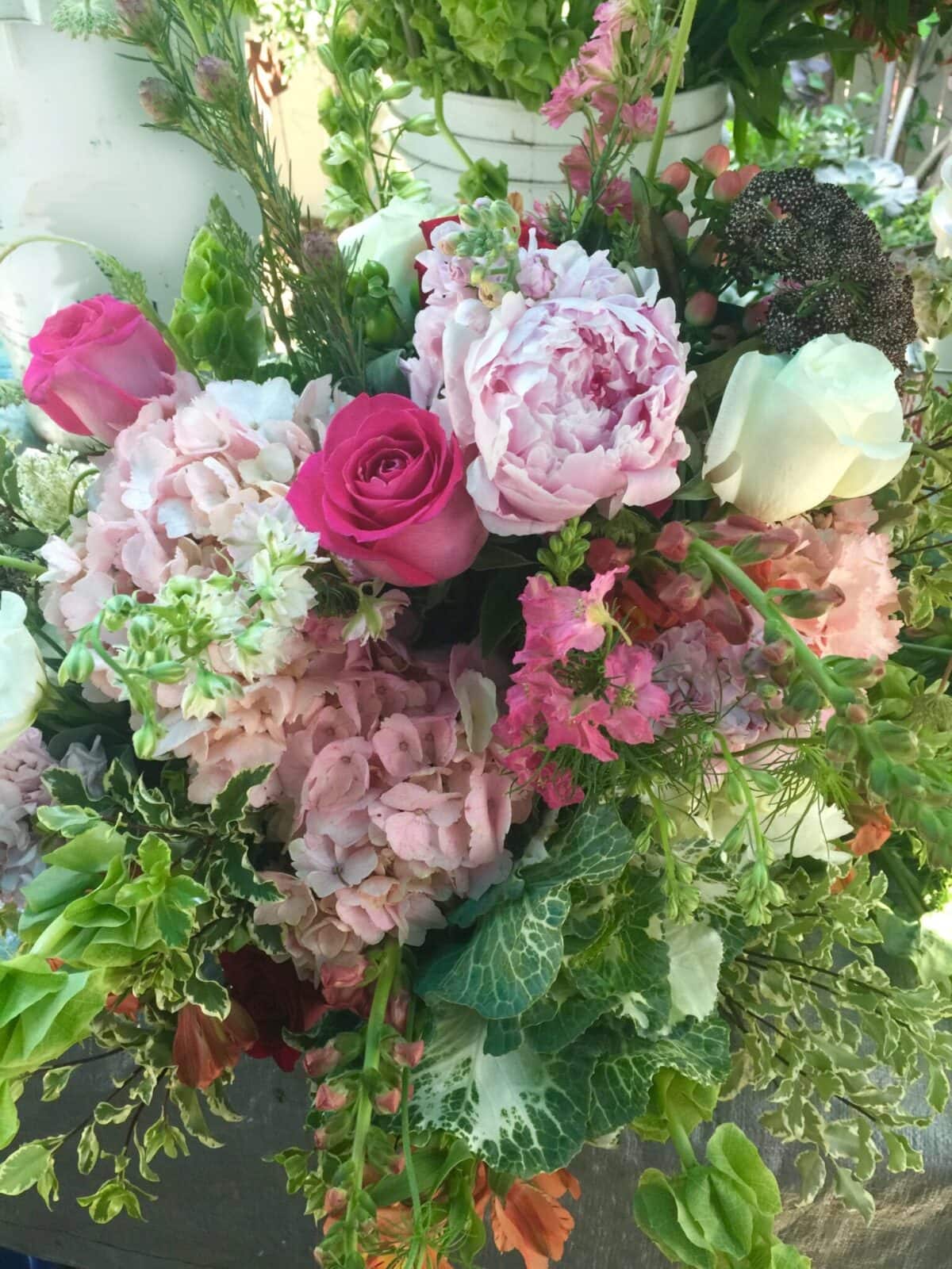 A colorful flower arrangement featuring pink and white roses, peonies, hydrangeas, stock flowers, and various green foliage in a lush and vibrant display—perfect for outdoor table centerpiece ideas.