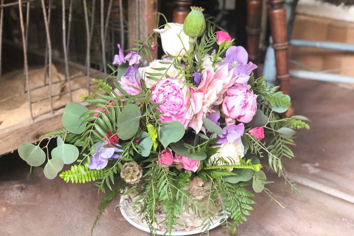 A lush floral arrangement featuring a variety of flowers including pink peonies, purple flowers, and white roses, complemented by abundant green foliage, is beautifully displayed in a rustic vase on a wooden surface.