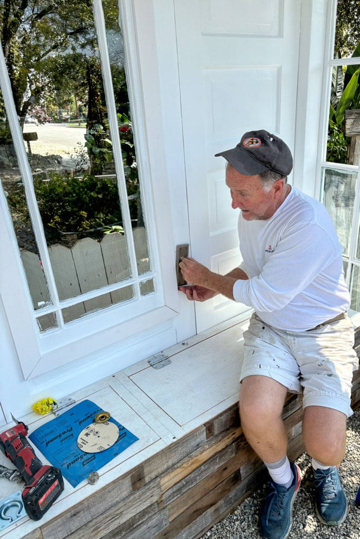 A man in a white shirt and shorts works on a window frame. He is sitting on a wooden platform holding a tool. Nearby are a red drill, a blue manual, and a tape measure. The background features greenery and another house.