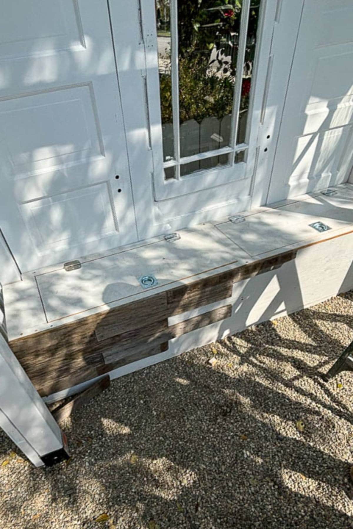 A white wooden bench with a backrest made from repurposed doors sits on a gravel surface. The bench has storage compartments indicated by hinges on the seats. There’s a tall mirror in the center backrest door, with some scattered shadows on the scene from nearby foliage.